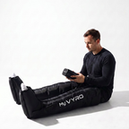 A man sits on a white studio floor wearing a black MyVYRO inflatable compression boots, holding a black handheld controller. The product is shown in use on a clean white background.
