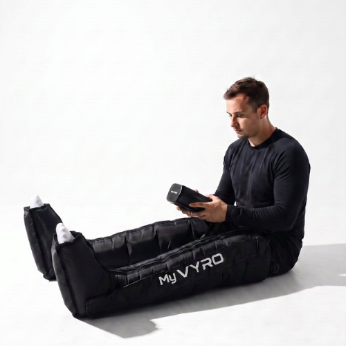 A man sits on a white studio floor wearing a black MyVYRO inflatable compression boots, holding a black handheld controller. The product is shown in use on a clean white background.
