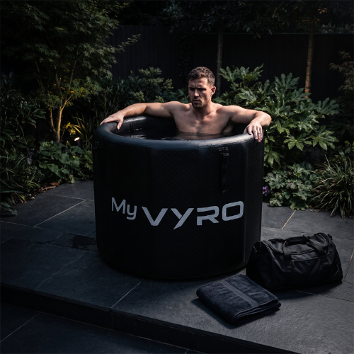 A man sits inside a black MyVYRO inflatable ice bath outdoors on a dark stone patio with greenery in the background. A black bag and folded towel are placed beside the tub.